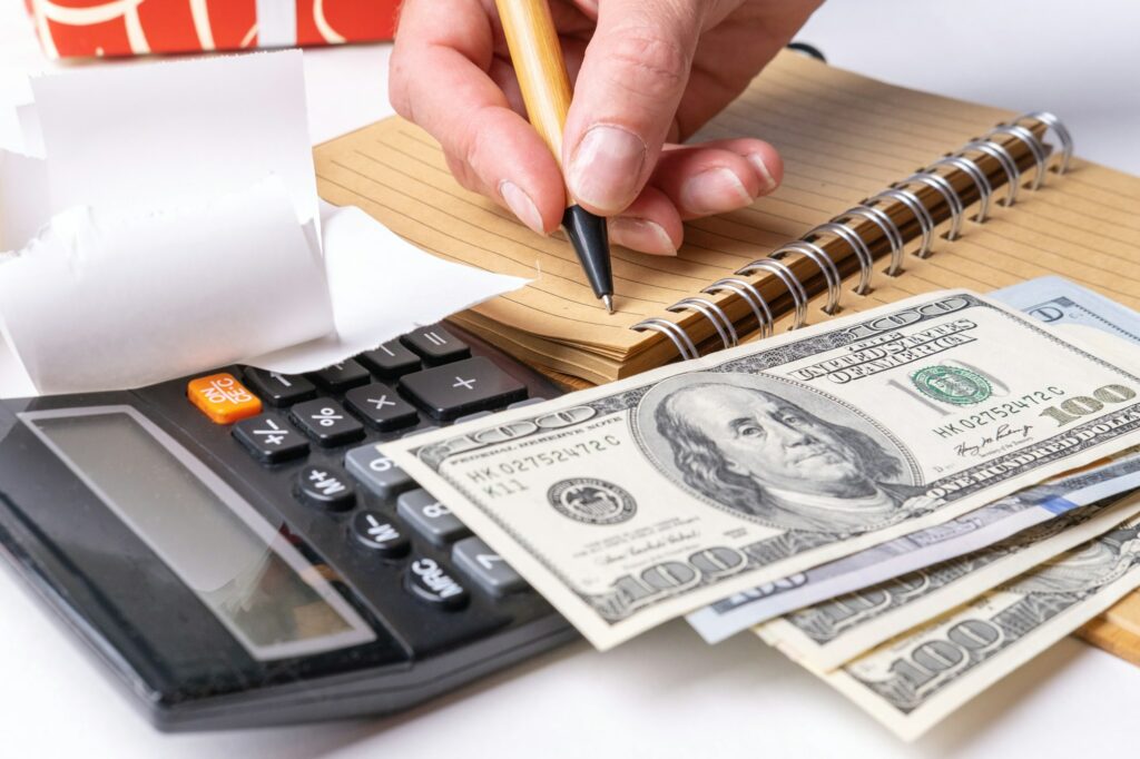 A woman's hand writing down the cash flow in a notebook with a pen, counting cash on a calculator